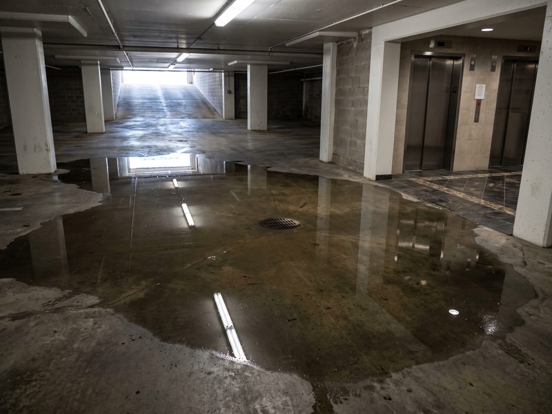 Shallow water across a below-grade parking level running toward a drain beside a high-rise elevator lobby.