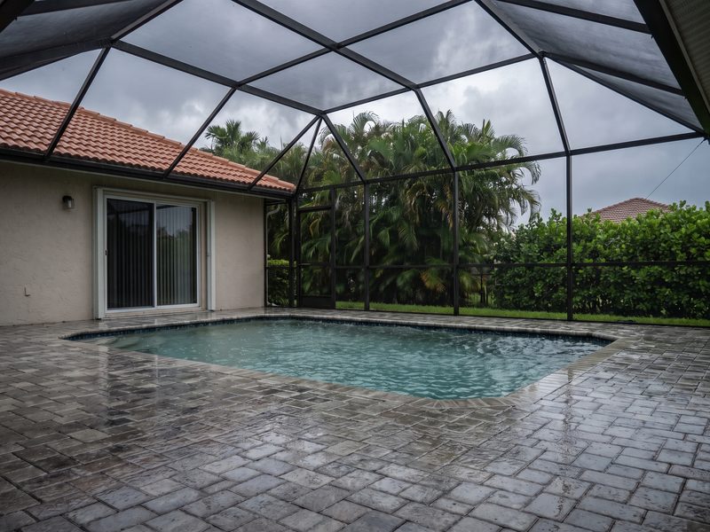 Rain falling into a screened pool enclosure, the pool near capacity and the paver deck standing wet beside the sliding door.