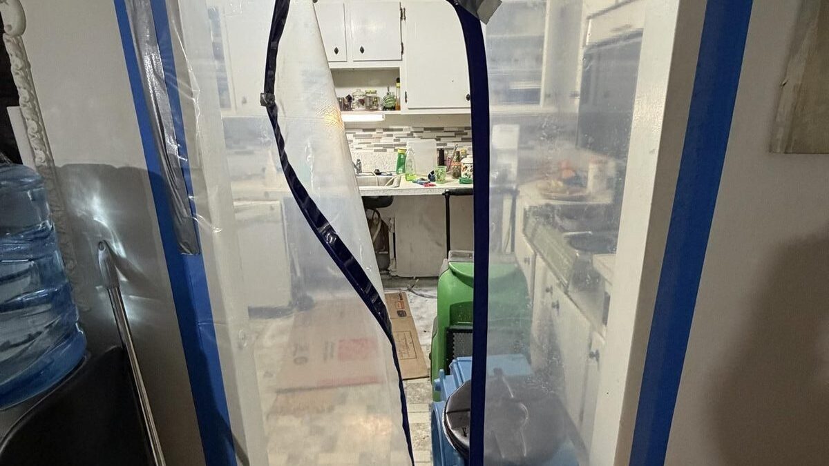 A zippered plastic containment barrier taped across a kitchen doorway with drying equipment behind it
