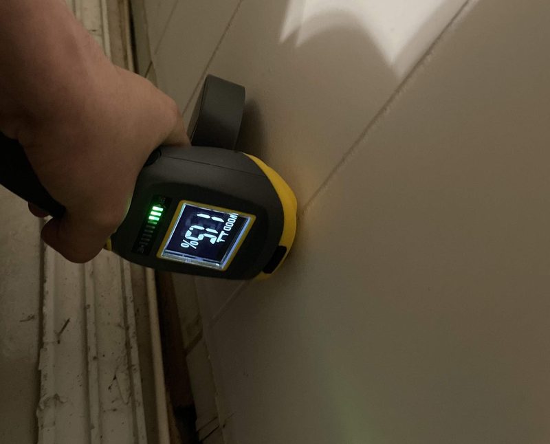 A hand holding a moisture meter flat against a wall at floor level in Hollywood, FL on October 25, 2024, its display turned sideways and reading 11.5 percent on the WOOD scale, Restoration Doctor project documentation.