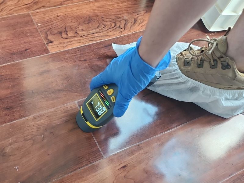 Crew member's boot in a white shoe cover beside a pinless moisture meter held to a wood-look plank floor in Kendall, FL, October 2024, Restoration Doctor project documentation.