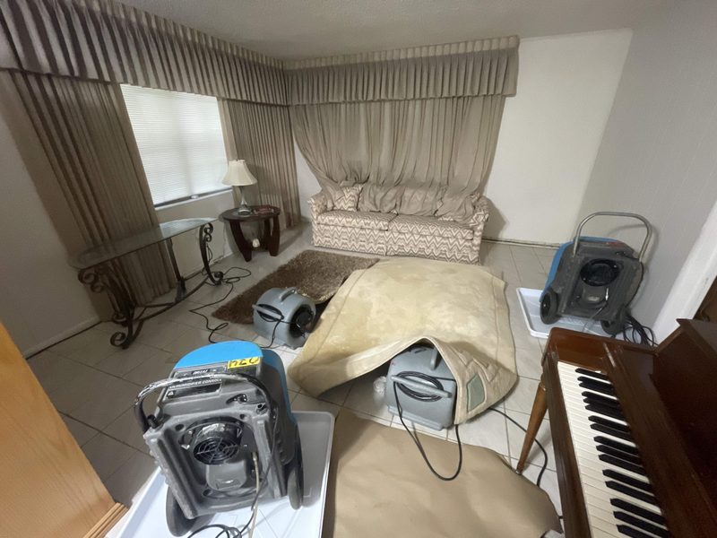 A living room in Hollywood, FL on October 25, 2024 with a cream rug lifted over an air mover, two dehumidifiers on drip trays and brown paper on the light tile floor, Restoration Doctor project documentation.