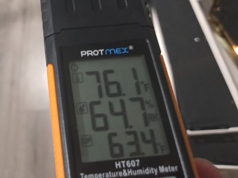A handheld temperature and humidity meter in a Miami Beach, FL unit on December 27, 2025, its display reading 76.1 degrees Fahrenheit and 64.7 percent relative humidity, Restoration Doctor project documentation.