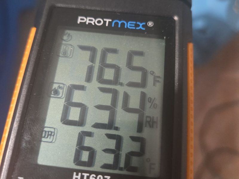 A handheld temperature and humidity meter in a Miami Beach, FL unit on January 6, 2026, its display reading 76.5 degrees Fahrenheit and 63.4 percent relative humidity, Restoration Doctor project documentation.