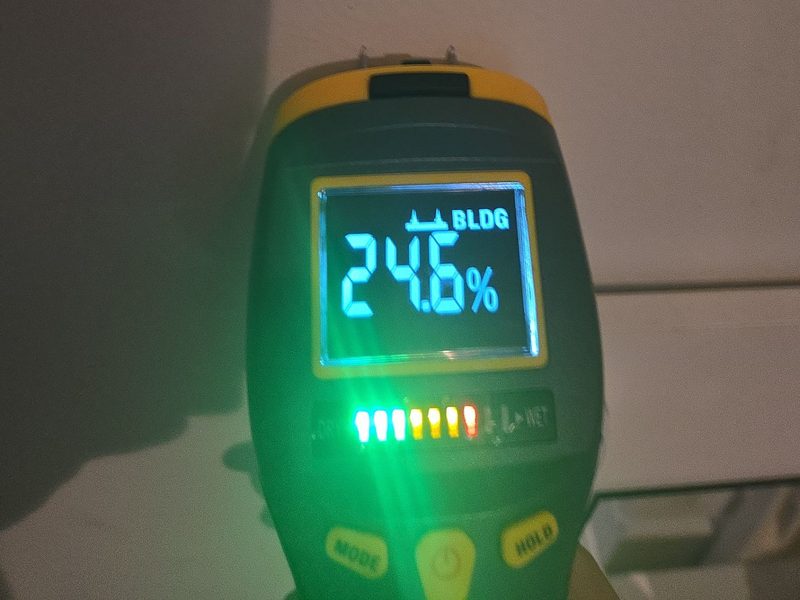 A pin-type moisture meter pressed to a wall in a Miami Beach, FL unit, December 2025, its display reading 24.6 percent on the BLDG scale, Restoration Doctor project documentation.