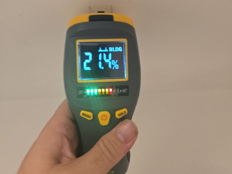 A hand holding a pin-type moisture meter against a wall in a Miami Beach, FL unit, December 2025, its display reading 21.4 percent on the BLDG scale, Restoration Doctor project documentation.